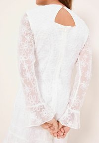 Valkoinen mekko, jossa kukkakirjailu, läpikuultavat pitkät hihat, avonainen selkä ja kellohihat. Pehmeä tekstuuri ja vetoketjusuljenta takana.