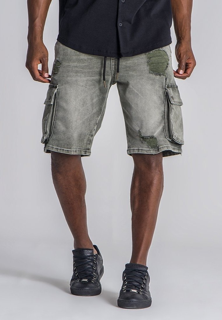 Bermudas en denim de color verde oliva deslavado, con detalles desgastados, múltiples bolsillos laterales y una cintura con cordón. Combinadas con zapatillas negras.