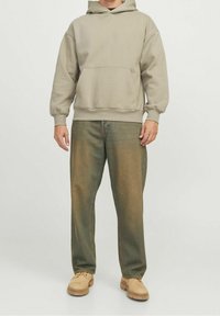 Sudadera beige con un bolsillo frontal, combinada con unos vaqueros de mezclilla verde. El calzado es unos zapatos casuales de color marrón claro.