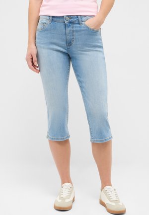 Femme portant un pantacourt en jean bleu clair et des baskets blanches, debout avec une main dans la poche.