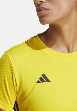 Gelbes Sportshirt mit rundem Ausschnitt. Verfügt über kurze Ärmel, ein strukturiertes Muster und ein dunkles Logo in der oberen linken Ecke.