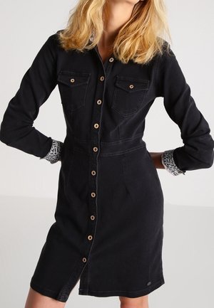 Kobieta ubrana w czarną, zapinaną na guziki sukienkę z denimu z długimi rękawami podwiniętymi, odsłaniającymi wzorzyste mankiety.