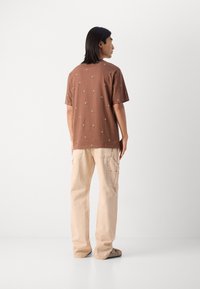 Brun bomulds t-shirt med små broderede blomster mønstre, parret med beige cargo bukser. Model set bagfra. Glat stof tekstur.