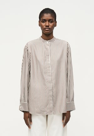 Isik kannab pruuni ja valge vertikaalsete triipudega pika varrukaga särk koos paelkraega ja valgeid pükse, seisab ühevärvilise tausta ees.