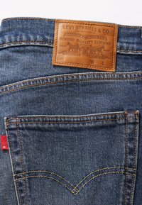 Jeans de mezclilla azul con una parche de cuero marrón en la pretina, presentando costuras anaranjadas y una etiqueta roja en el bolsillo trasero.