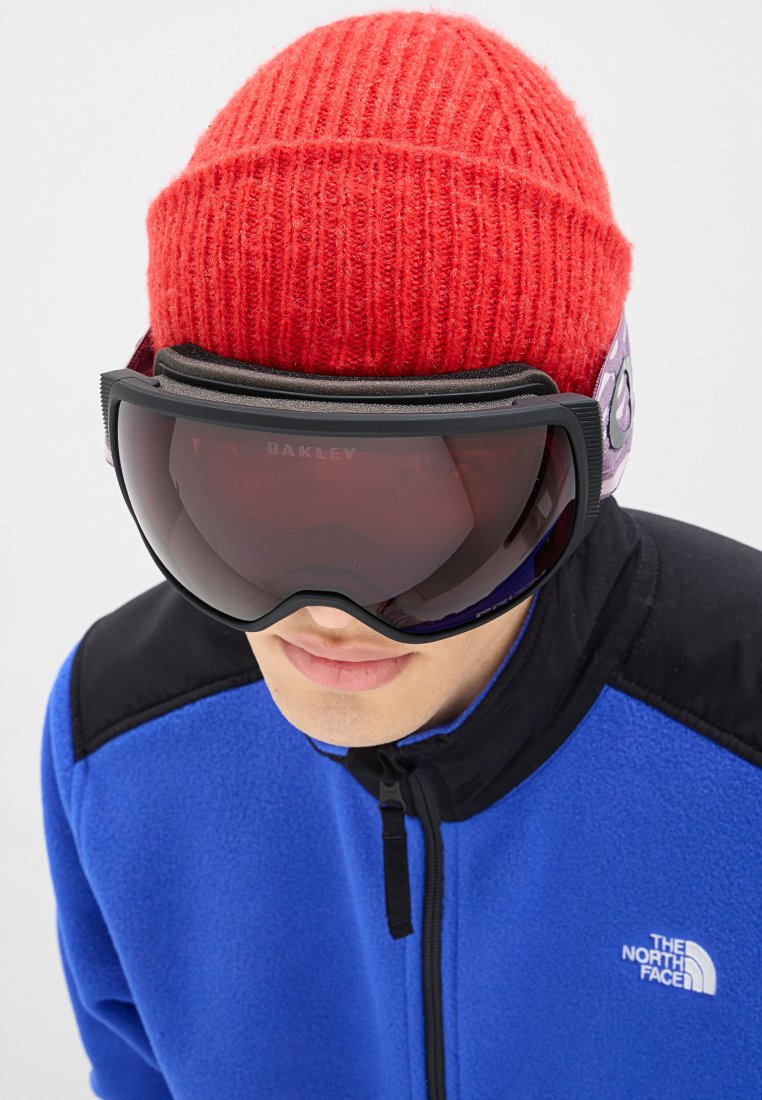 Osoba носząca duże czarne gogle narciarskie, czerwony wełniany czapkę oraz niebieską i czarną kurtkę North Face na jednolitym tle.
