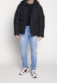 Chaqueta de plumas negra con capucha, cierres de presión en el frente y dos grandes bolsillos laterales, combinada con jeans de color azul claro y zapatillas multicolores.