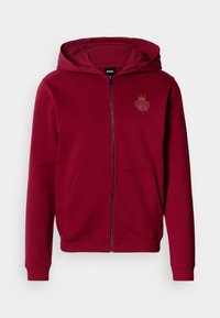 Červená bavlnená mikina na zips s klokankovým vreckom a kapucňou so šnúrkami. Má malé reliéfne logo na ľavej strane hrude. Hladká textúra.