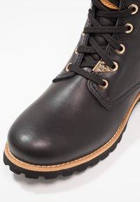 Botte en cuir noir à bout arrondi, surface texturée, œillets dorés, lacets noirs, et semelle extérieure contrastante couleur tan avec coutures visibles.