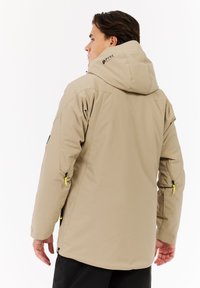 Beige Outdoor-Jacke mit Kapuze, ausgestattet mit Reißverschlusstaschen, schwarzen Akzenten und kontrastierenden gelben Reißverschlüssen. Glattes, wetterfestes Material.