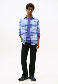 Camicia a quadri blu chiaro con accenti bianchi e rossi, indossata sopra un maglione viola a maglia, abbinata a pantaloni neri e scarpe da ginnastica nere.