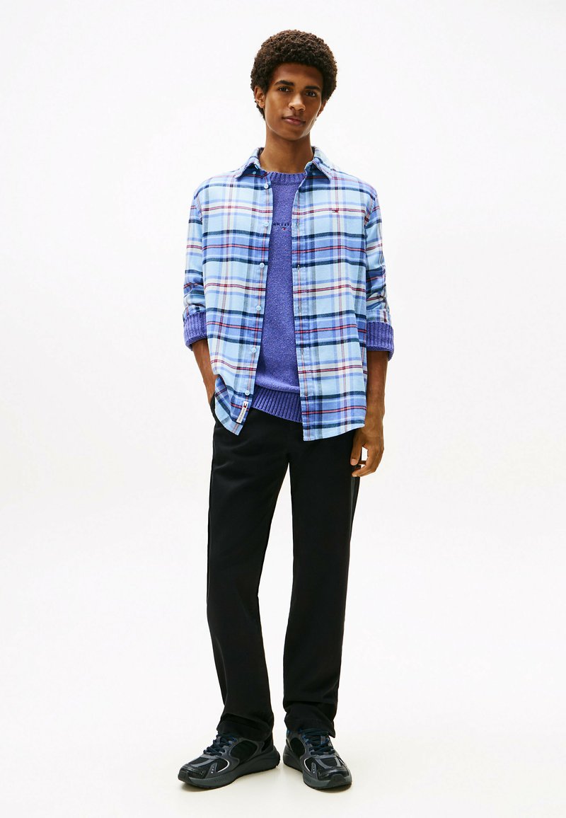 Chemise à carreaux bleu clair avec des accents blancs et rouges, superposée sur un pull en tricot violet, associée à un pantalon noir et des chaussures de sport noires.