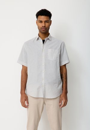 Mann, der vor weißem Hintergrund steht, trägt ein hellgraues kurzärmeliges Hemd und beige Hose, mit einem Tattoo am linken Unterarm.