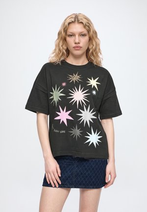 Un mannequin aux cheveux blonds ondulés porte un T-shirt noir orné d’imprimés colorés en forme d’étoiles rayonnantes et une jupe en denim bleu foncé à motifs.