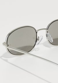 Lunettes de soleil ovales avec des verres gris, monture en métal argenté, plaquettes de nez transparentes et branches fines. Reflets de lumière visibles sur les verres.