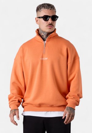 Sweat-shirt en polaire orange avec un col montant et une demi-fermeture éclair. Présente des poignets et un ourlet côtelés, avec le logo "ESTEEM" en blanc sur la poitrine.