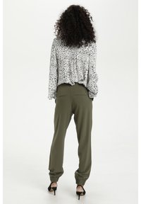 Weißes Blusenshirt mit schwarzen Sprenkeln, lockerer Schnitt. Olivgrüne Jogginghose mit elastischen Bündchen und Gesäßtaschen. Schwarze High-Heel-Sandalen.