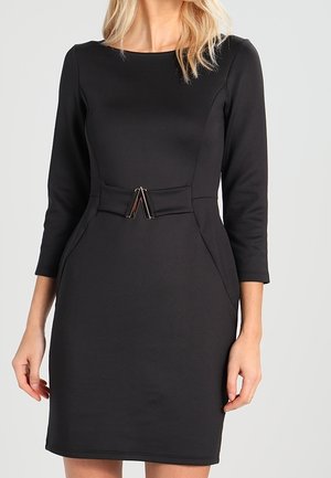 Frau trägt ein eng anliegendes schwarzes knielanges Kleid mit dreiviertel Ärmeln und einem dekorativen Gürtel mit einer Metallschnalle in der Taille.