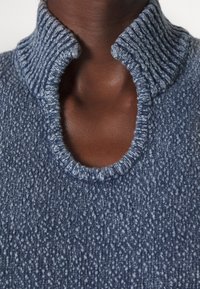 Gros plan d'une personne portant un pull en maille bleue texturée avec un col montant côtelé déplié et un décolleté ouvert.