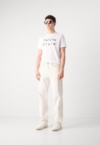 White cotton t-shirt with black "CALVIN KLEIN" text, paired with cream-colored jeans and white sneakers. Model wearing dark sunglasses.