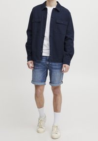 Veste-chemise marine avec boutons sur le devant et deux poches poitrine, associée à un short en denim bleu retroussé et des baskets blanches à semelles texturées.