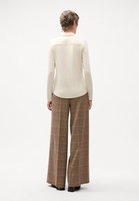 Beige Bluse mit langen Ärmeln und einer rückwärtigen Naht, kombiniert mit braunen weiten Karohosen mit einem karierten Muster und seitlichen Taschen.