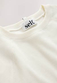 Gros plan sur le col d’une chemise en tissu blanc côtelé avec une étiquette de vêtement blanche indiquant « self. » cousue à l’intérieur du col.