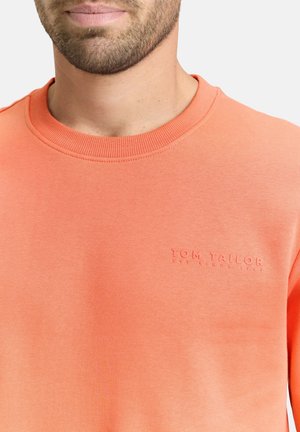 Mann trägt einen korallenfarbenen Pullover mit Rundhalsausschnitt und eingeprägtem "Tom Tailor"-Logo auf der Brust, mit leichtem Bart und neutralem Hintergrund.
