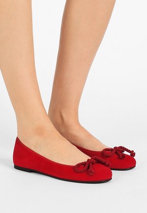 Beine mit roten Ballettschuhen mit abgerundeten Zehen und großen dekorativen Schleifen aus Seil obenauf, vor einem schlichten weißen Hintergrund.