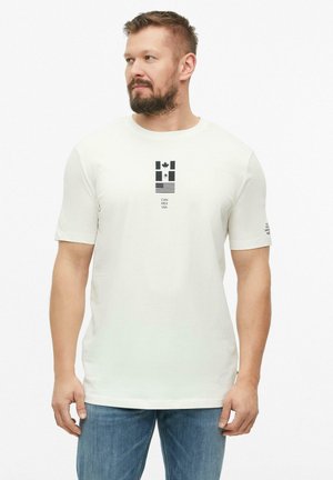 Man met baard die een wit T-shirt draagt met de vlaggen van Canada, Mexico en de VS en tekst, gecombineerd met blauwe spijkerbroek, kijkt naar links tegen een witte achtergrond.