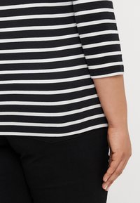 Czarna i biała blouse w paski z rękawami do łokcia, wykonana z miękkiego, gładkiego materiału. Widoczne ręce i czarne spodnie z detalem kieszeni.