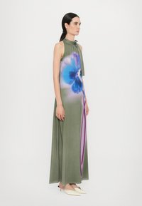 Lange, mouwloze jurk in olijfgroen met een groot blauw bloemenpatroon en paarse accenten. Bevat een strik halslijn en vloeiende stof.