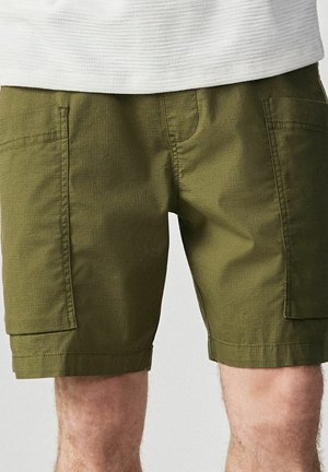 Olivgrüne cargoförmige Shorts in Knielänge mit aufgesetzten Taschen vorne, kombiniert mit einem weißen, strukturierten Oberteil.