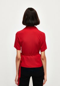 Blouse rouge à manches courtes avec un col et une ceinture à nouer à la taille, confectionnée dans un tissu lisse et léger.