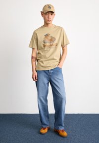 Beige cotton t-shirt with a graphic print, paired with relaxed blue denim jeans. Tan sneakers with maroon accents complete the outfit.