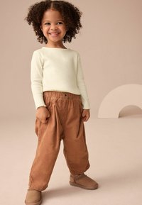 Maglietta a maniche lunghe a coste verde chiaro, pantaloni ampi in tessuto morbido marrone con vita elasticizzata e scarpe beige slip-on su uno sfondo neutro.
