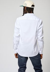 Witte lange mouwen knoopshirt met een soepele textuur, met een gebogen zoom en geknoopte manchetten. G gedragen met donkerblauwe jeans.