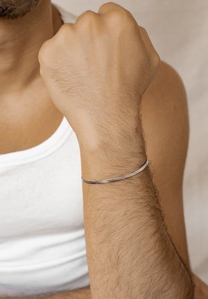 Male arm raised with clenched fist, wearing a thin silver bracelet and a white sleeveless shirt against a plain background.