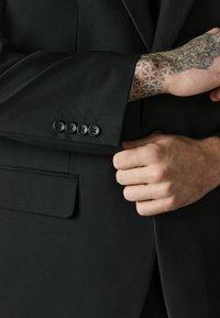Main tatouée ajustant le poignet d'une veste de costume noire avec quatre boutons noirs et une poche à rabat.