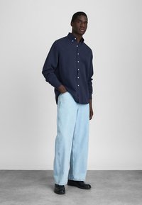 Un homme se tenant avec une main dans la poche, portant une chemise bleu marine à boutons, un jean large bleu clair et des chaussures noires sur un sol gris.