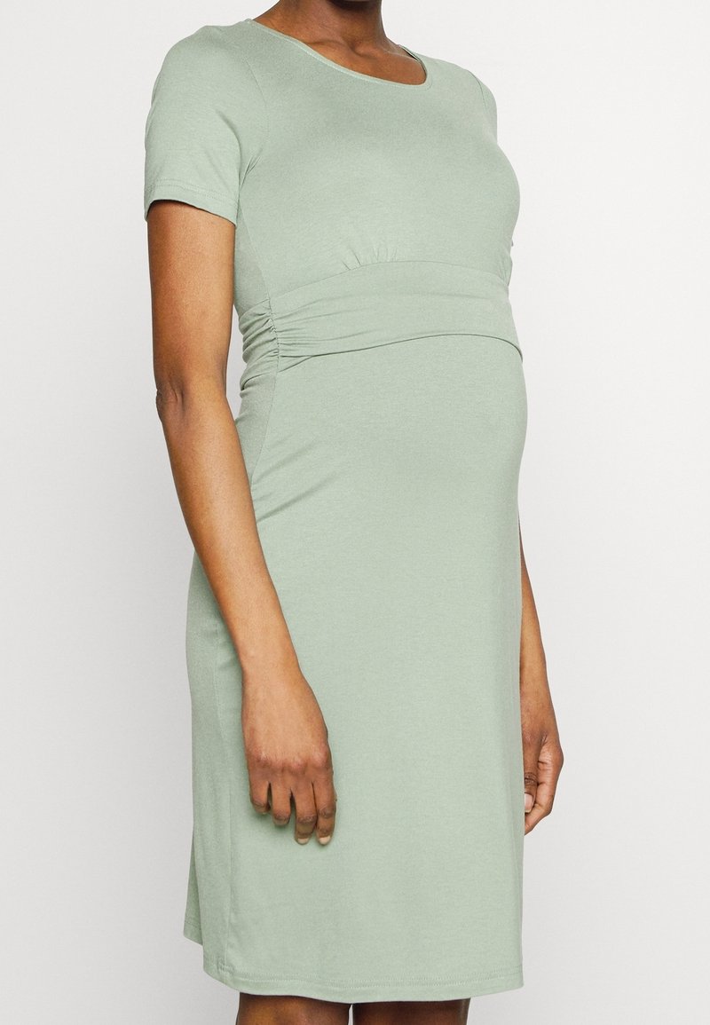 Personne enceinte portant une robe de maternité vert clair, à manches courtes, longueur genoux, avec des côtés froncés et un décolleté arrondi.