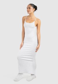 Kvinde med håret sat op i en knold iført en tætsiddende lysegrå ærmeløs maxi-kjole og hvide sneakers, stående mod en ensfarvet lys baggrund.