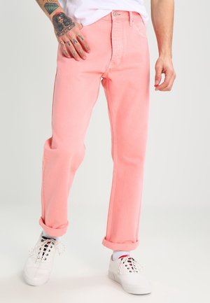Uomo con pantaloni rosa risvoltati, sneakers bianche, calzini bianchi e camicia bianca, con tatuaggi visibili sull'avambraccio sinistro e sulla mano.