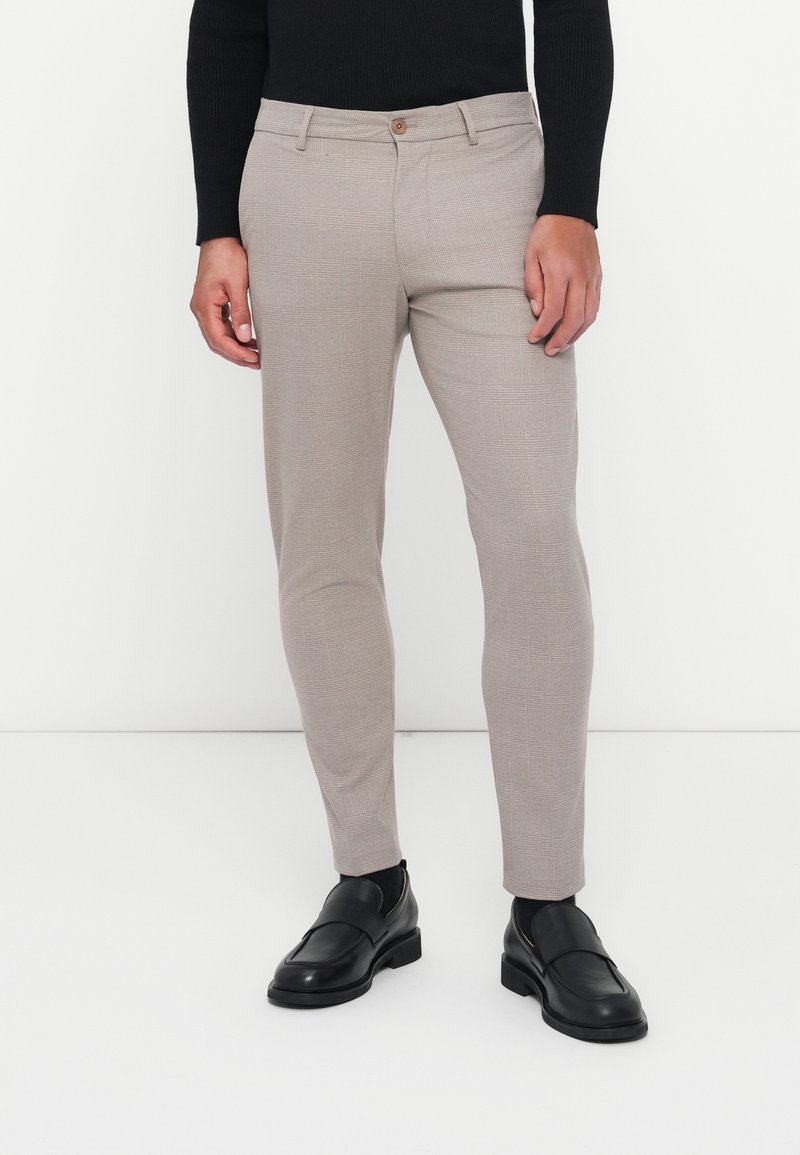Beige, skreddersydde bukser med et subtilt rutemønster, slim fit, og en enkel brun knapp i midjen. Kombinert med svarte loafers.