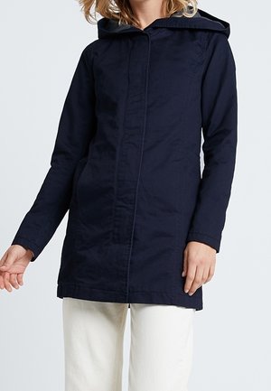 Femme portant une longue veste à capuche bleu marine avec une fermeture avant cachée et un pantalon blanc, debout devant un fond uni.