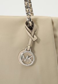 Beige mulepose med tekstureret stof, der har et håndtag i sølvkæde og en rund logomærkat fastgjort med en læderrem.