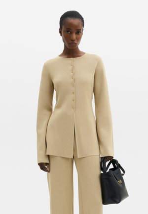 Femme portant un haut beige à manches longues boutonné et un pantalon assorti, tenant un sac à main noir avec des détails dorés sur un fond blanc uni.