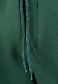 Mørkegrønt stof med flettede snørebånd, der hænger ned og viser tekstur og syningsdetaljer.