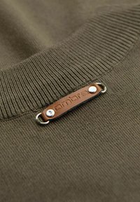 Pull en tricot vert olive avec un col côtelé, agrémenté d'une étiquette en cuir marron avec un texte en relief et des boutons pression en métal argenté.