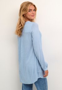 Žena s dlouhými blond vlasy, oblečená v světle modré blůze s dlouhým rukávem a modrých džínách, dívající se přes rameno na bílém pozadí.